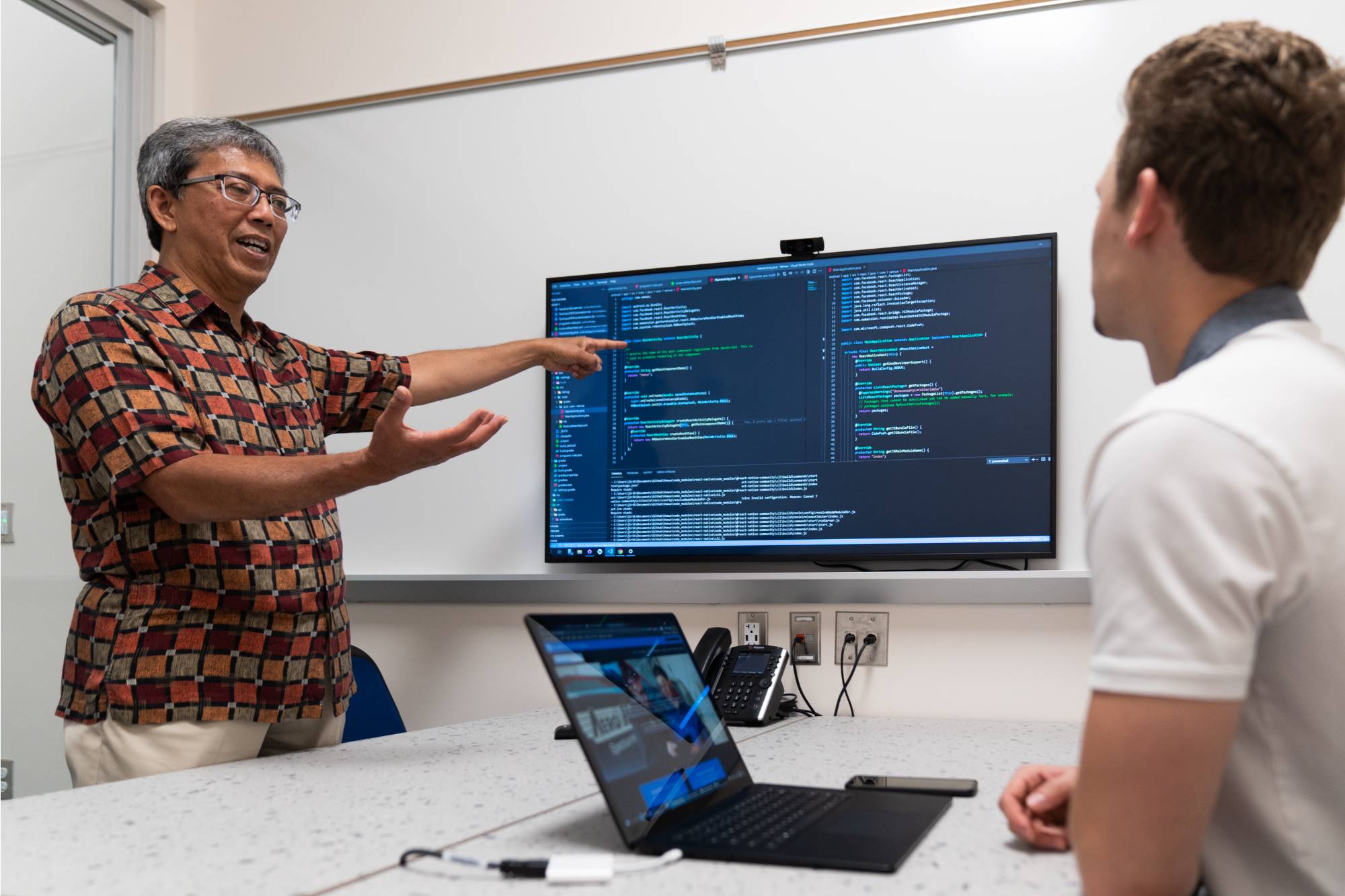 two people at a table talking with a large flatscreen displaying computer coding